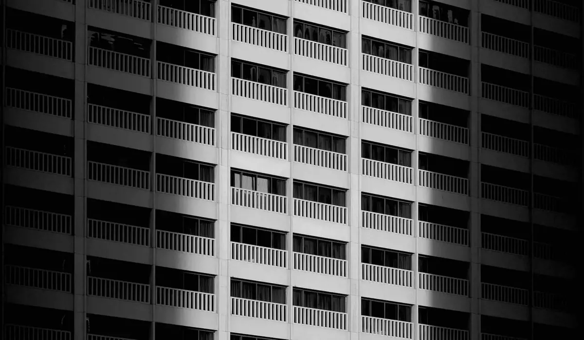 Black-and-white photo of a tall apartment building facade with rows of identical balconies, half in sunlight and half in shadow