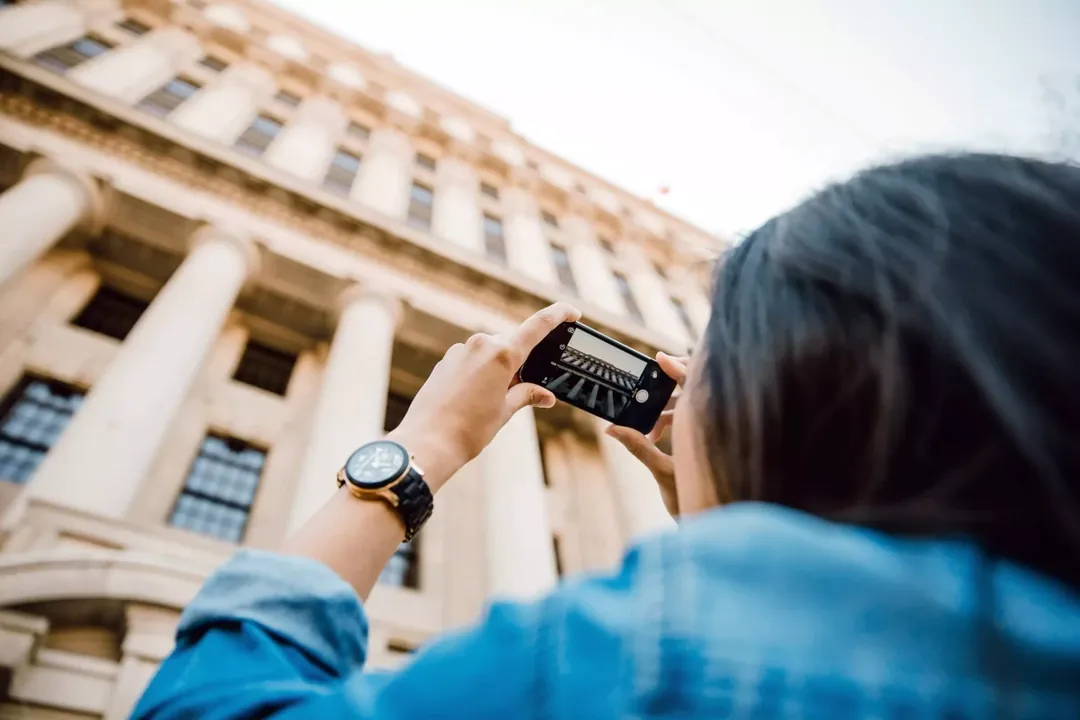 Woman with dark hair taking a photo with her phone of a large building with stone columns