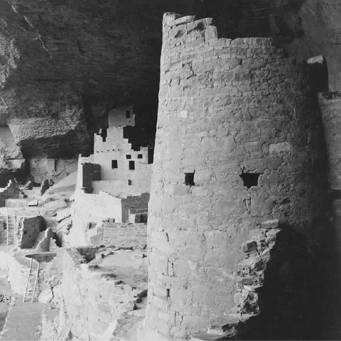 Black and white photo of a round stone tower and walls of the Cliff Palace cliff dwelling at Mesa Verde, built on a rock ledge