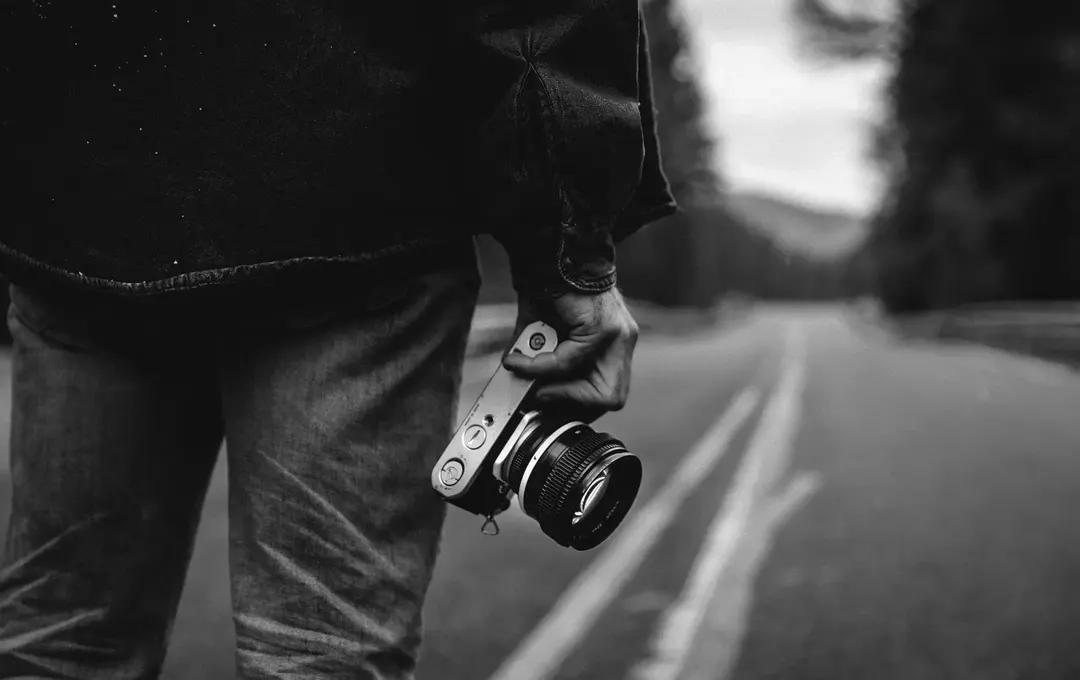 Black-and-white photo of a person standing on a road holding a camera at their side