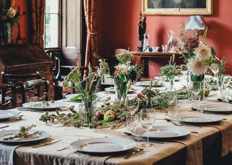A dining room with deep red walls crowded with antique furniture, a writing desk, vases, framed art and a long table covered in plates, glasses and dried flowers