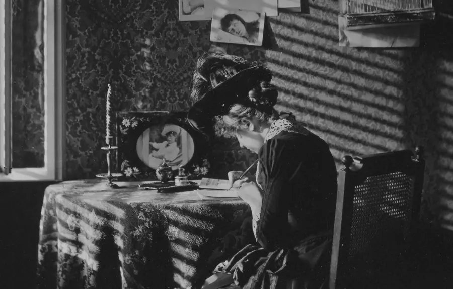 Vintage black and white photo of a woman writing at a desk covered with papers, a framed photo and a candlestick, in a room with patterned wallpaper