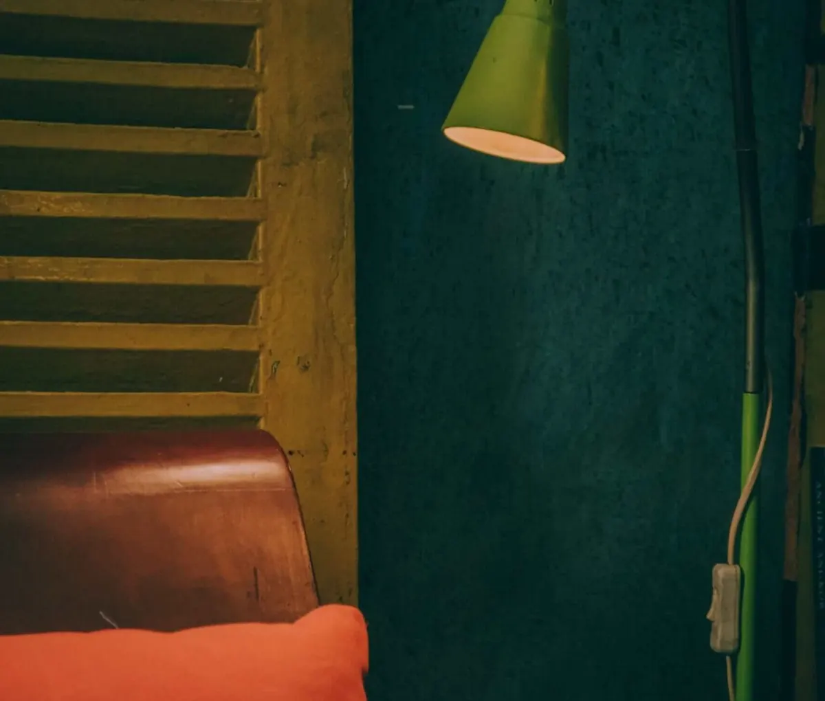 Corner of a lived-in room with a green desk lamp lighting a dark teal wall, the curved back of a leather armchair and an orange cushion in front of an old wooden shutter