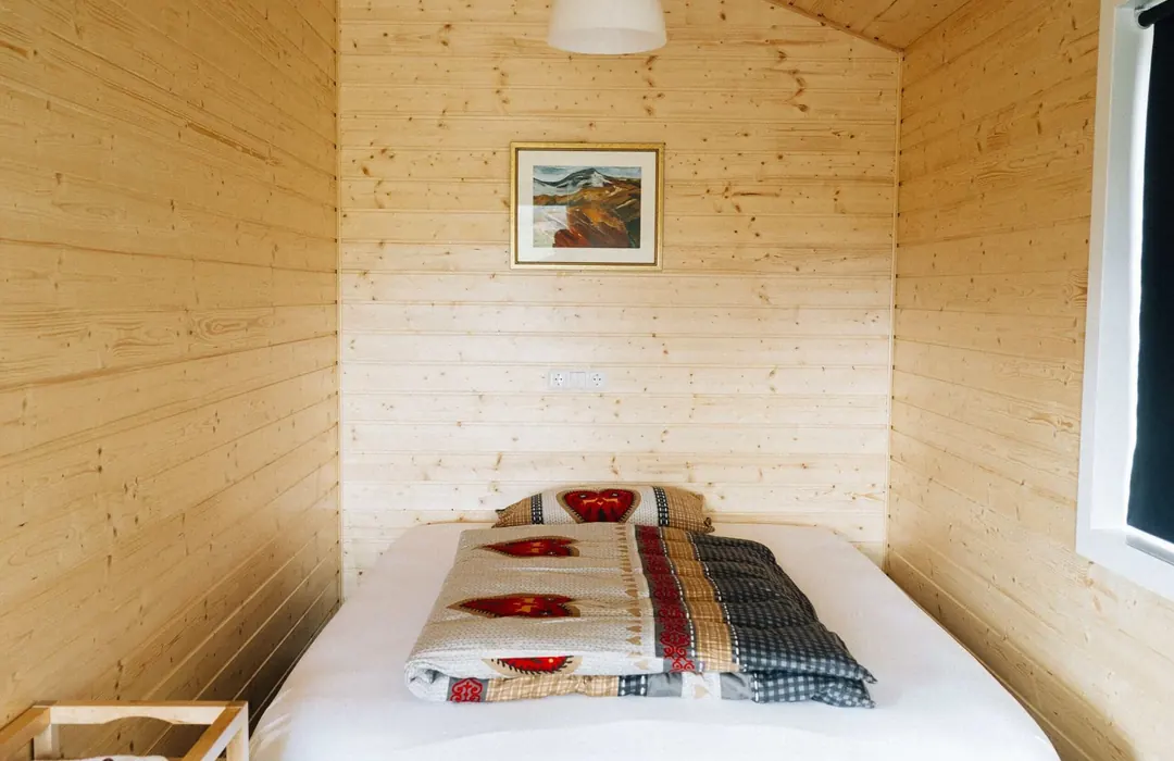 Small, simple bedroom with light pine walls, a neatly made bed with a patterned quilt, a framed painting and a bright window