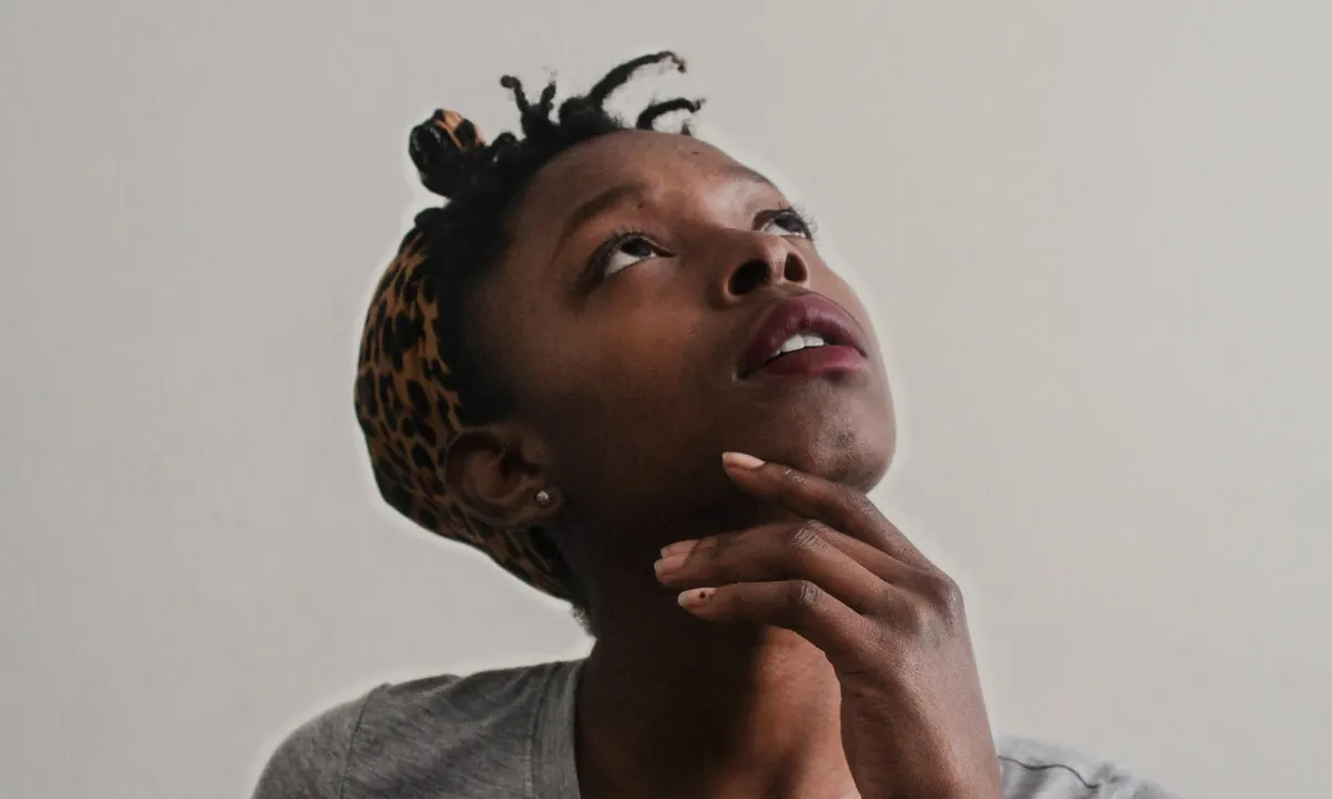 Young woman with her hand on her chin, looking up thoughtfully against a plain gray wall