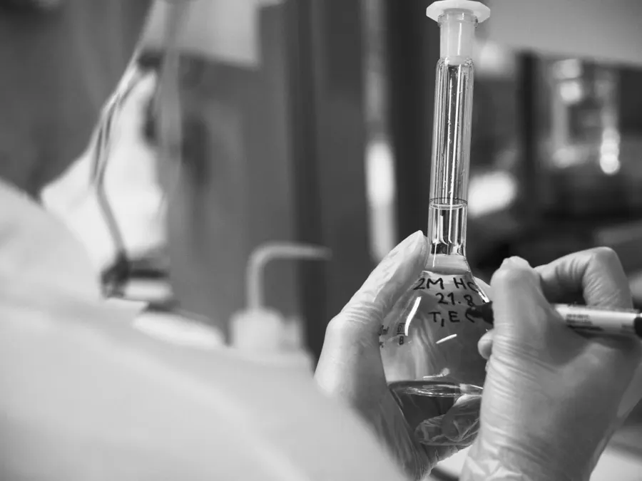 Black and white photo of a lab worker in gloves holding a glass flask and a pipette