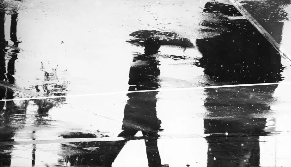Black and white photo of a person with an umbrella reflected in a rain-soaked street