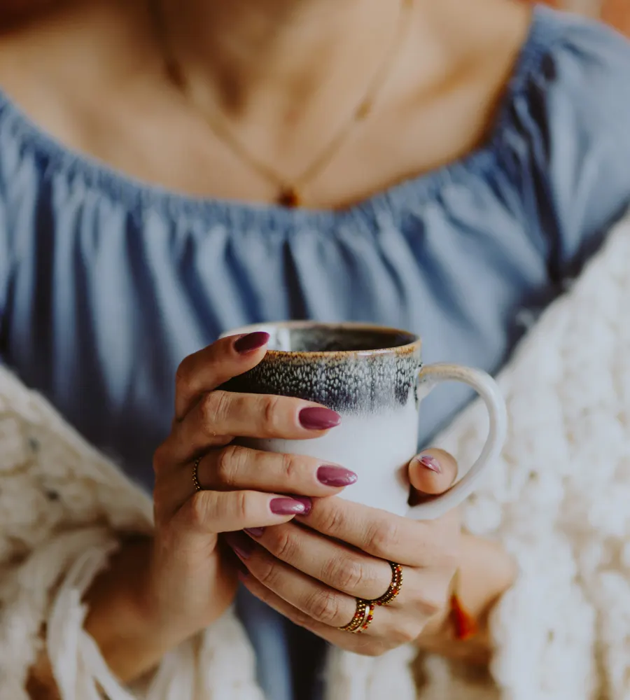 Woman wrapped in a cream knit blanket holding a ceramic mug with both hands