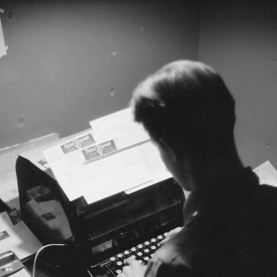 Black and white photo of a person seen from behind typing on an old typewriter, with stacks of forms and papers on the desk