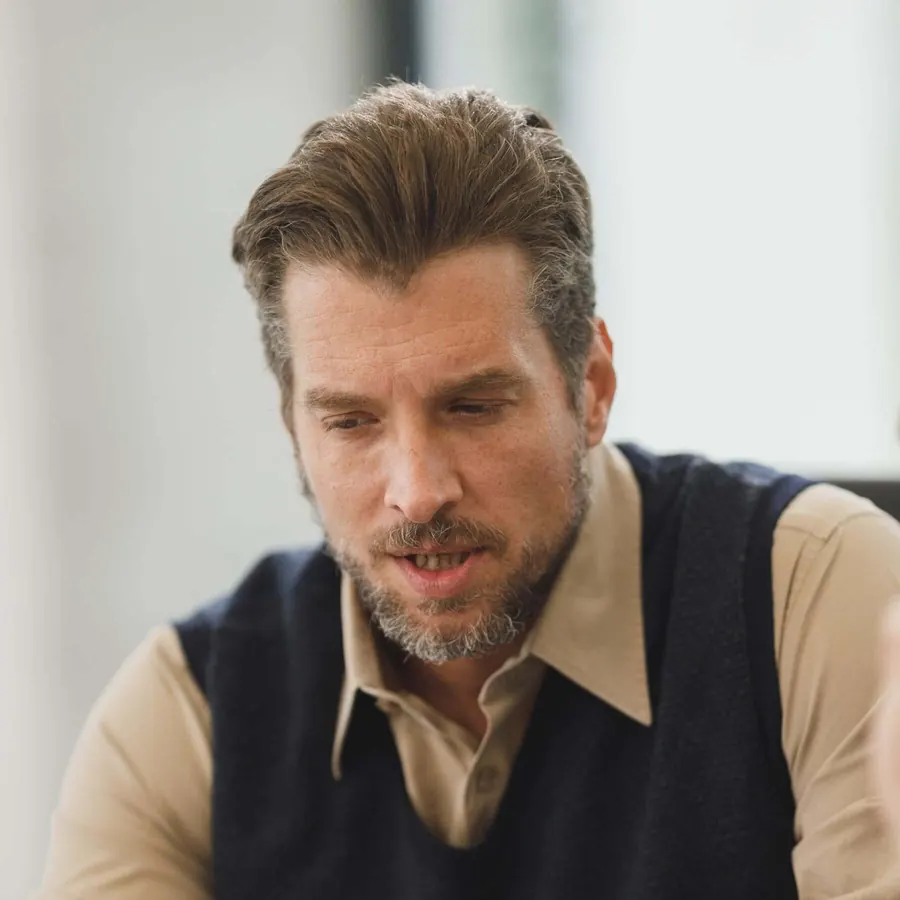Middle-aged man with a beard in a collared shirt and dark sweater vest, looking down thoughtfully while seated indoors