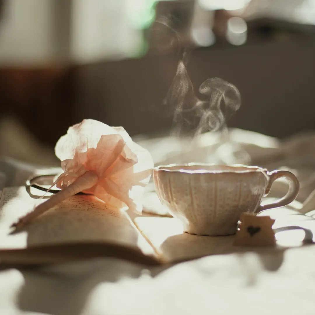 Close-up of a steaming white teacup with a pink flower resting on the pages of an open book, in soft warm light