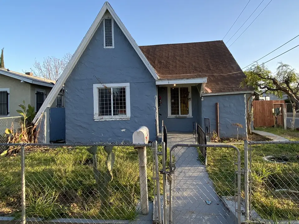 Small older blue cottage-style house with a steep gable roof behind a chain-link fence in the Fresno area